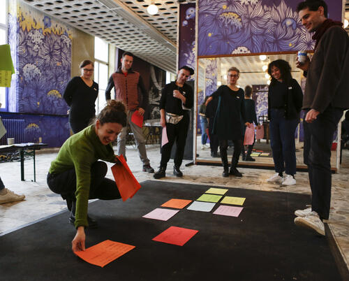 Copy Paste participants circle around a carpet on which are display sheets of colors with handwrittend sentences on them.